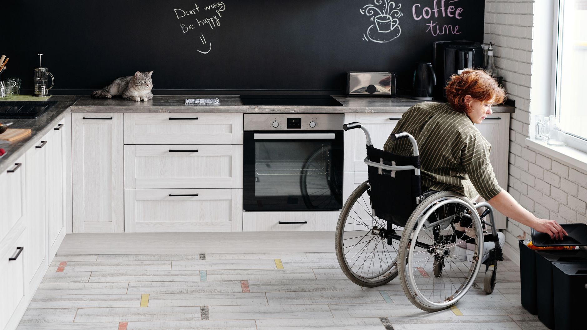 Woman in wheelchair using modern kitchen with cat and chalkboard wall. Warm and inviting atmosphere.