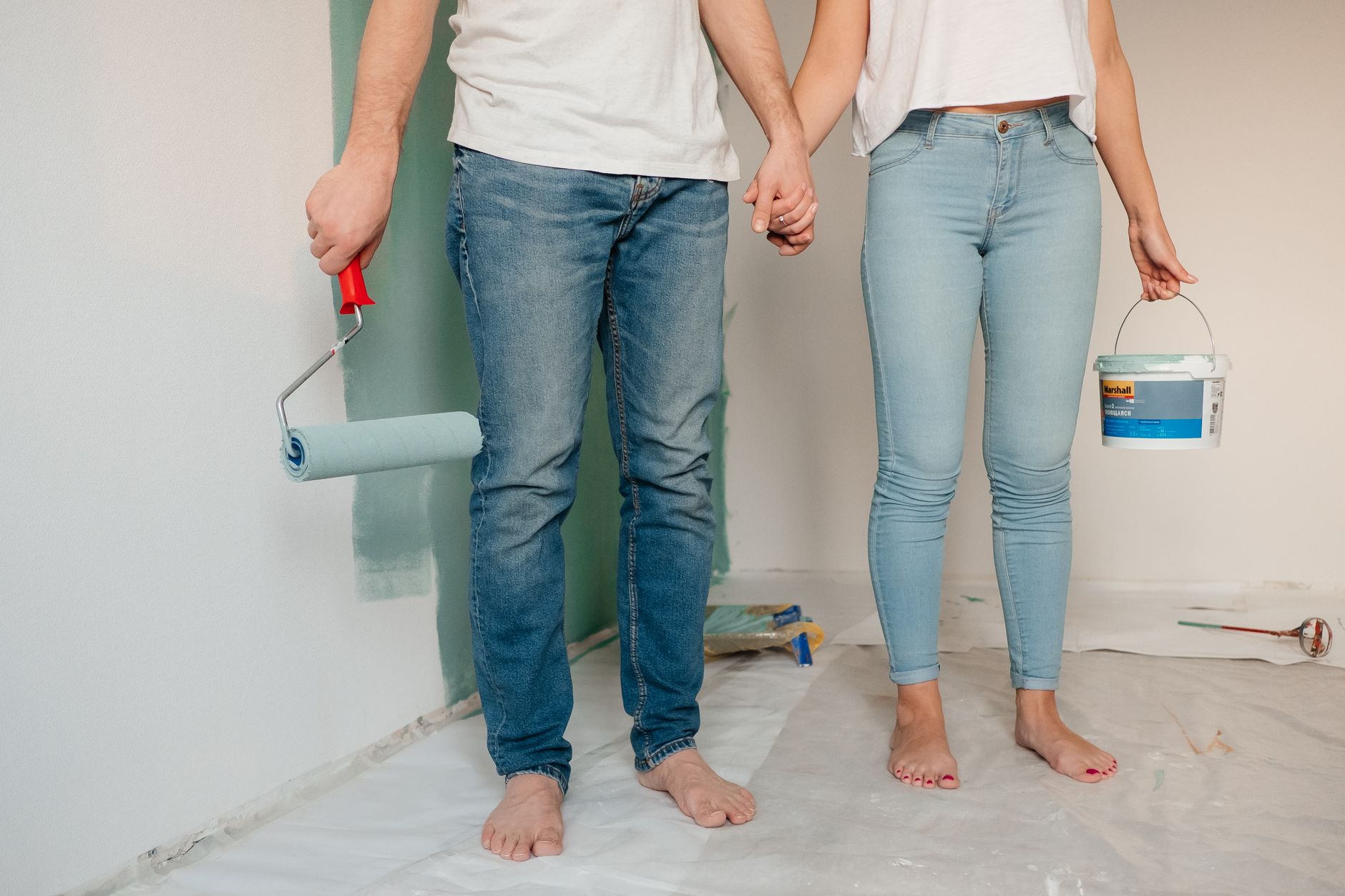 A couple engaged in home renovation, painting a wall barefoot and holding hands, symbolizing love and teamwork.