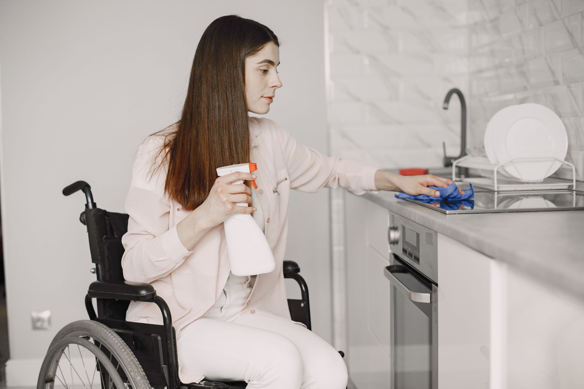 A woman in a wheelchair cleaning a modern kitchen countertop using a spray bottle and cloth.