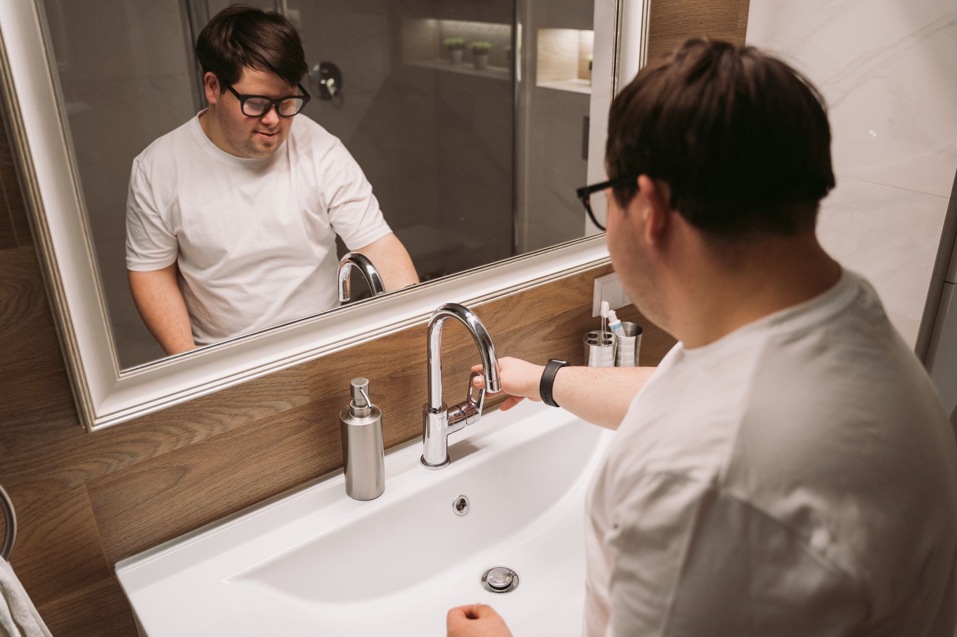 A man with Down syndrome using a bathroom sink while wearing glasses.