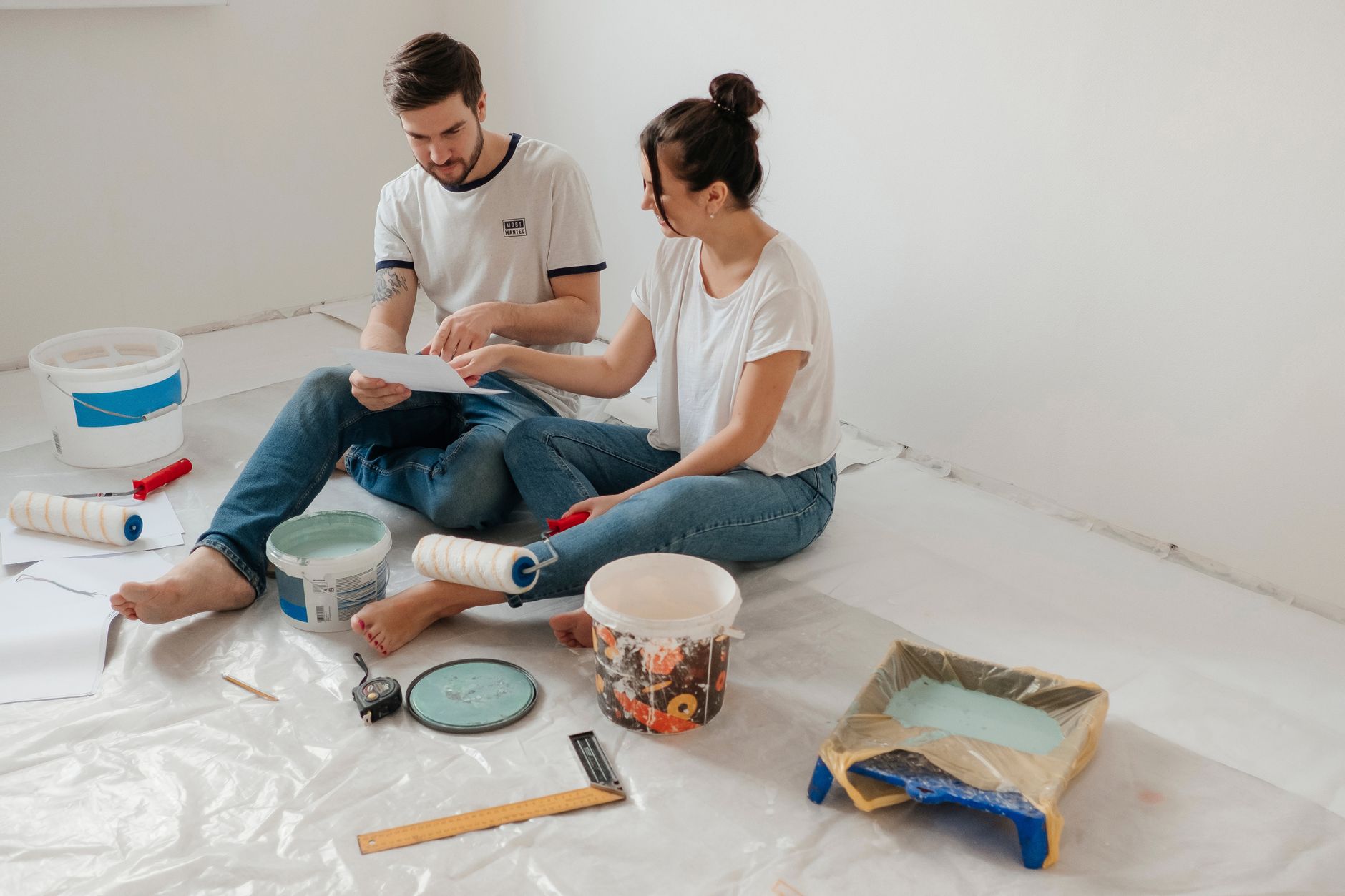A young couple discusses renovation plans while sitting on the floor surrounded by painting supplies.