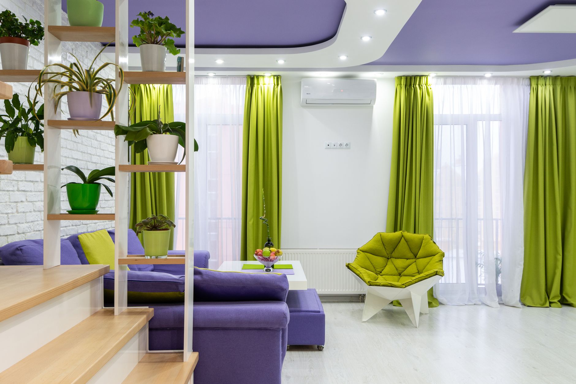 Interior of modern living room with bright purple sofa and ceiling and green armchair and curtains decorated with potted plants placed on cr
