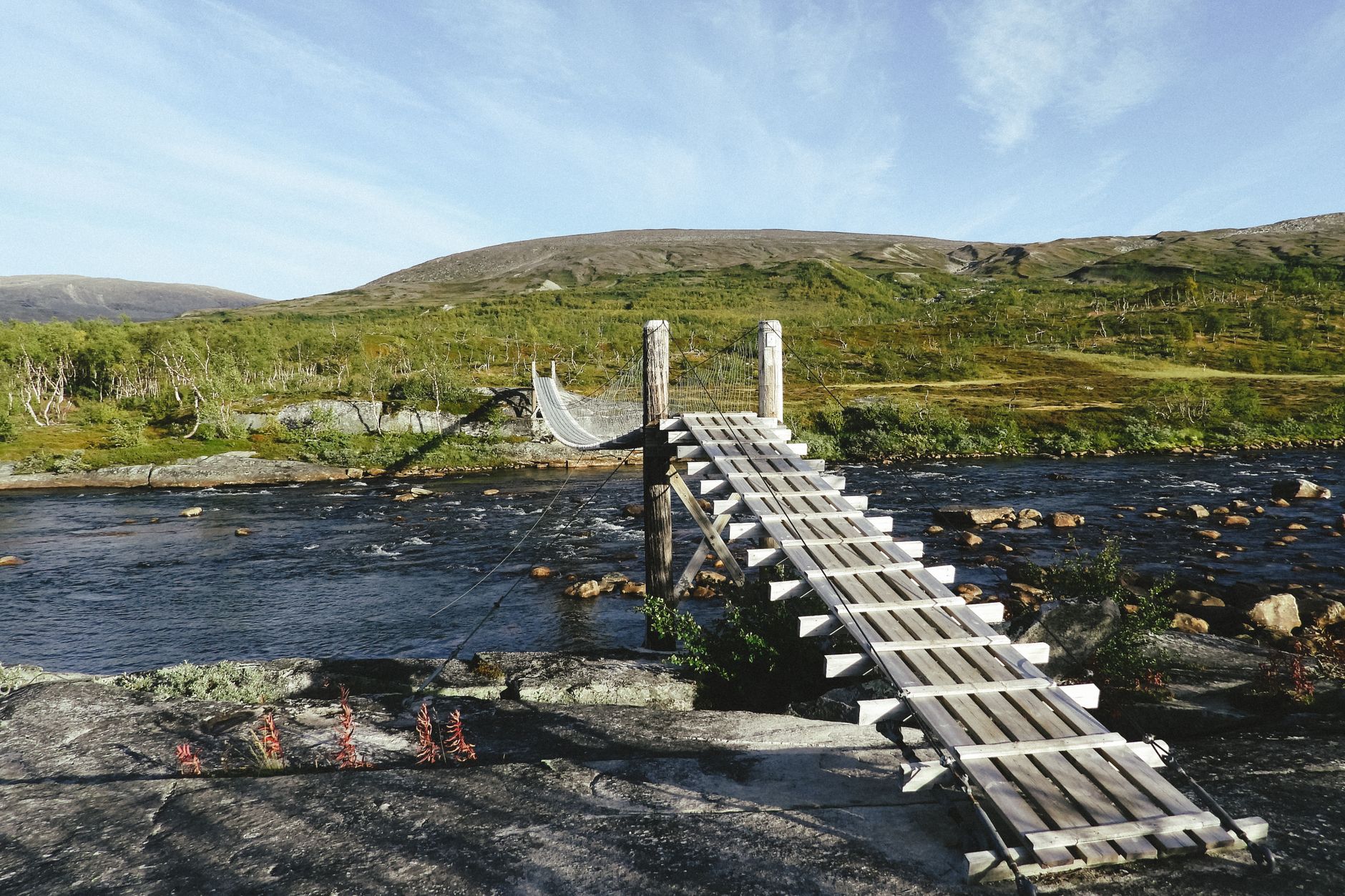 Wooden walkway over fast river flowing in green hilly terrain under blue sky
