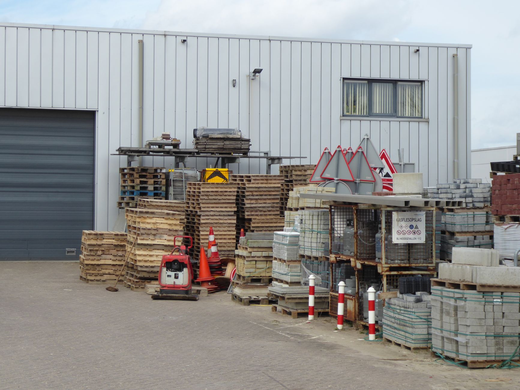 An outdoor yard with stacked construction supplies and pallets against a warehouse.