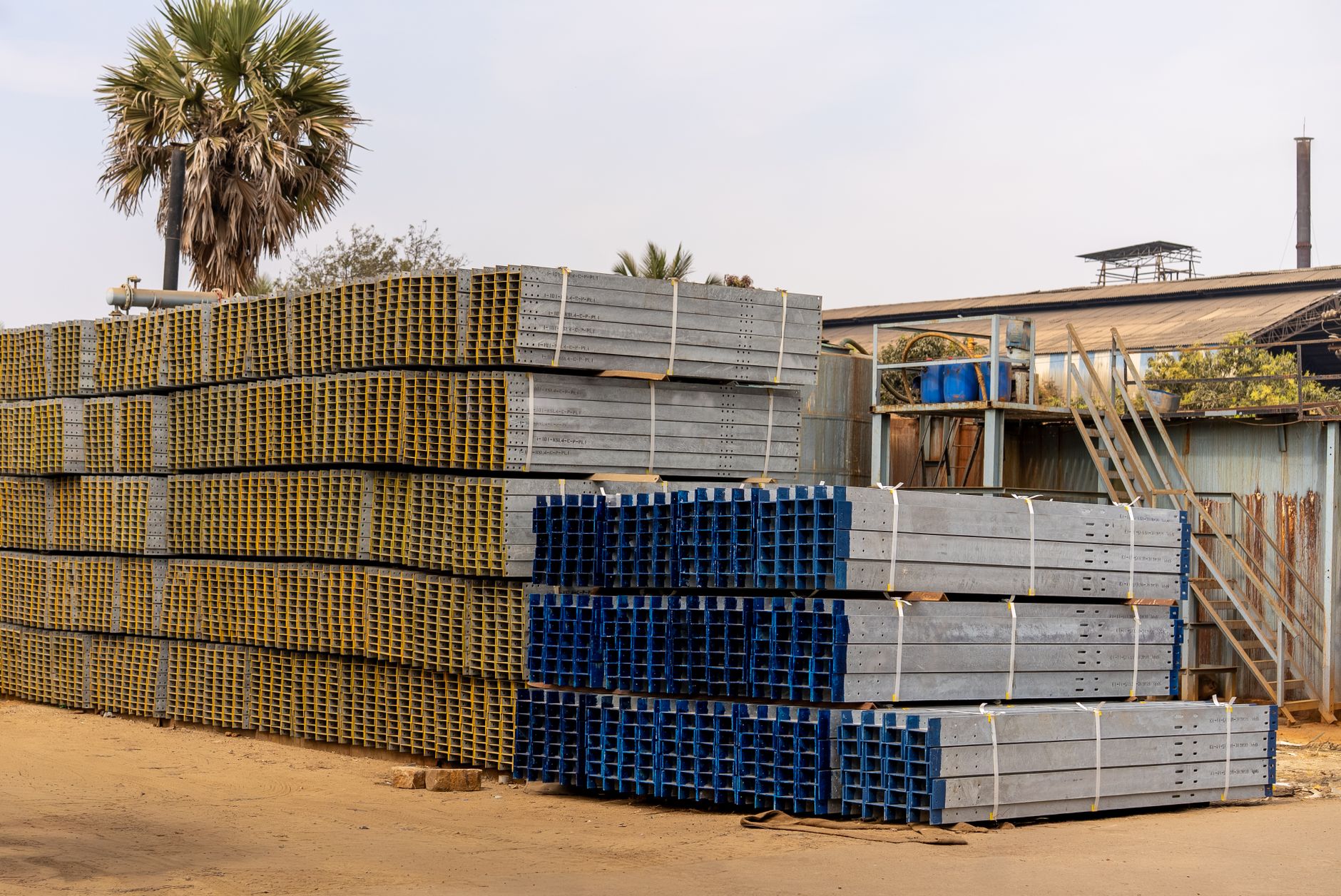 Piles of stacked metal beams in an industrial outdoor yard under a clear sky.